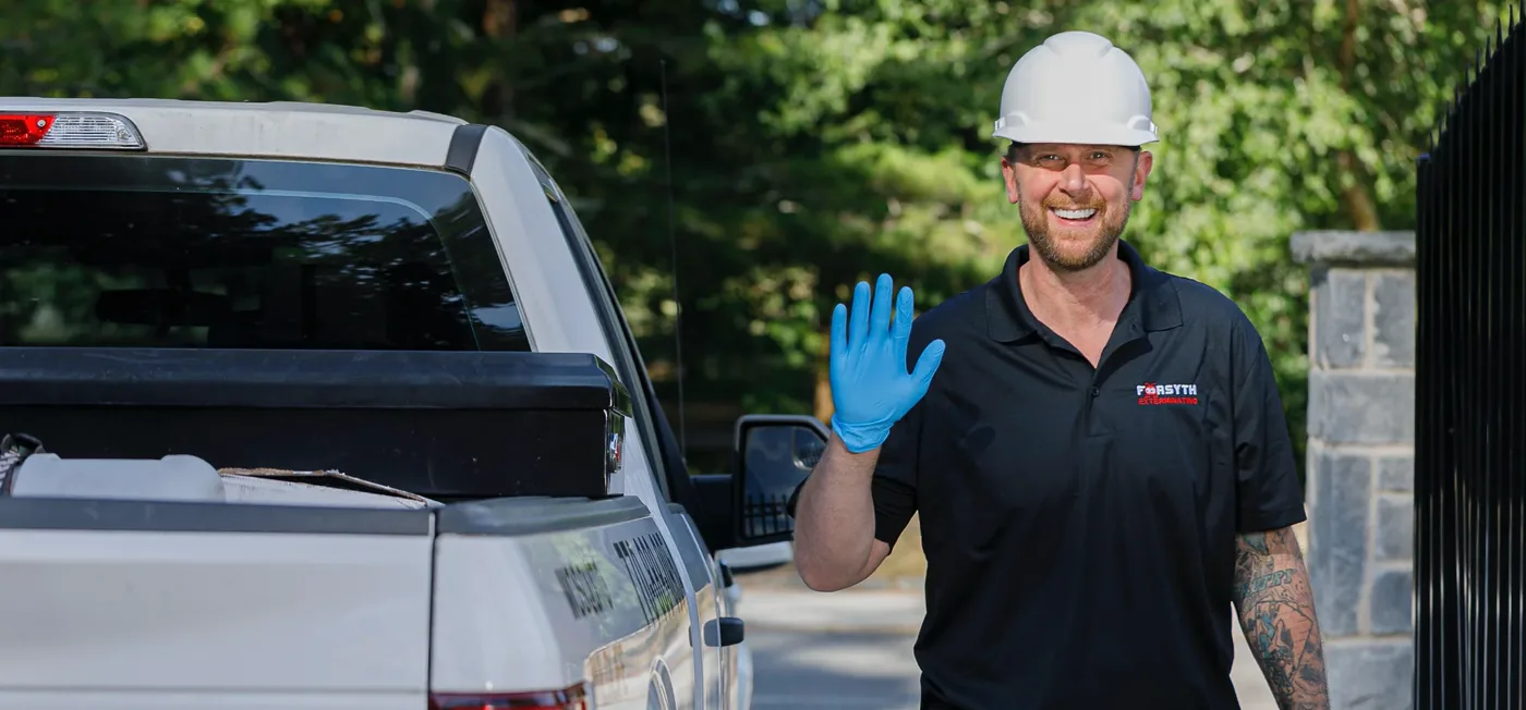 A Forsyth Exterminating technician in a hard hat waving beside a white car